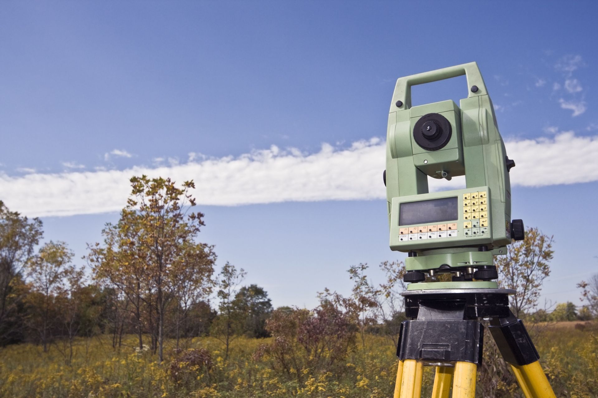 Surveying instrument on a tripod in a field, blue sky and trees in the background.
