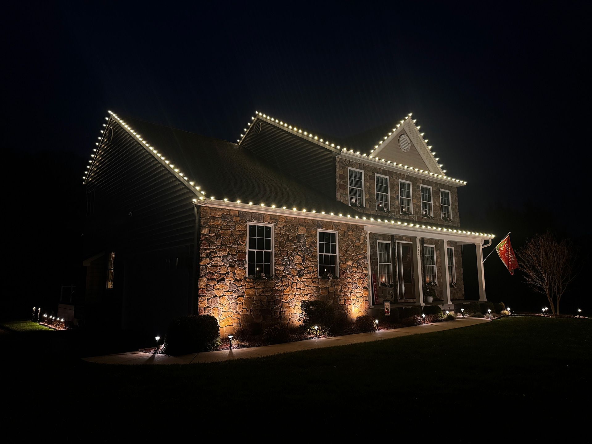 Two-story brick house illuminated with white Christmas lights along the roofline and ground-level spotlights.