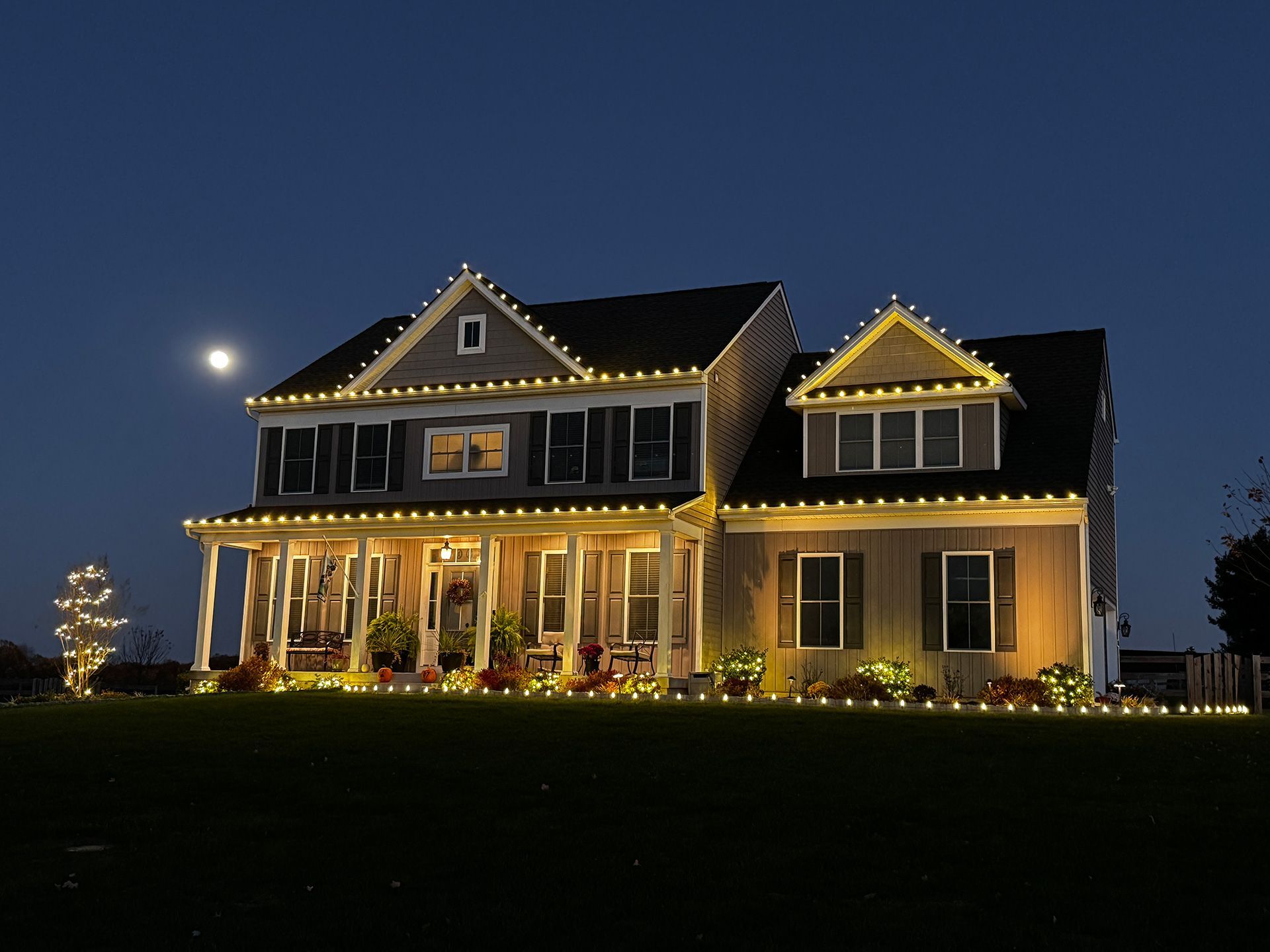 House lit with yellow Christmas lights at night, full moon in the sky.