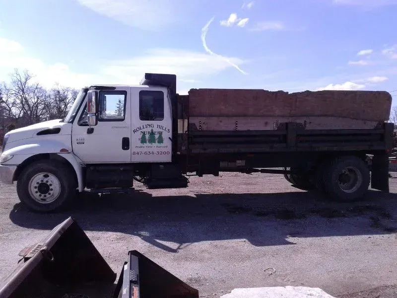 A white dump truck with a dark cargo bed parked on a gravel lot under a bright, clear blue sky.