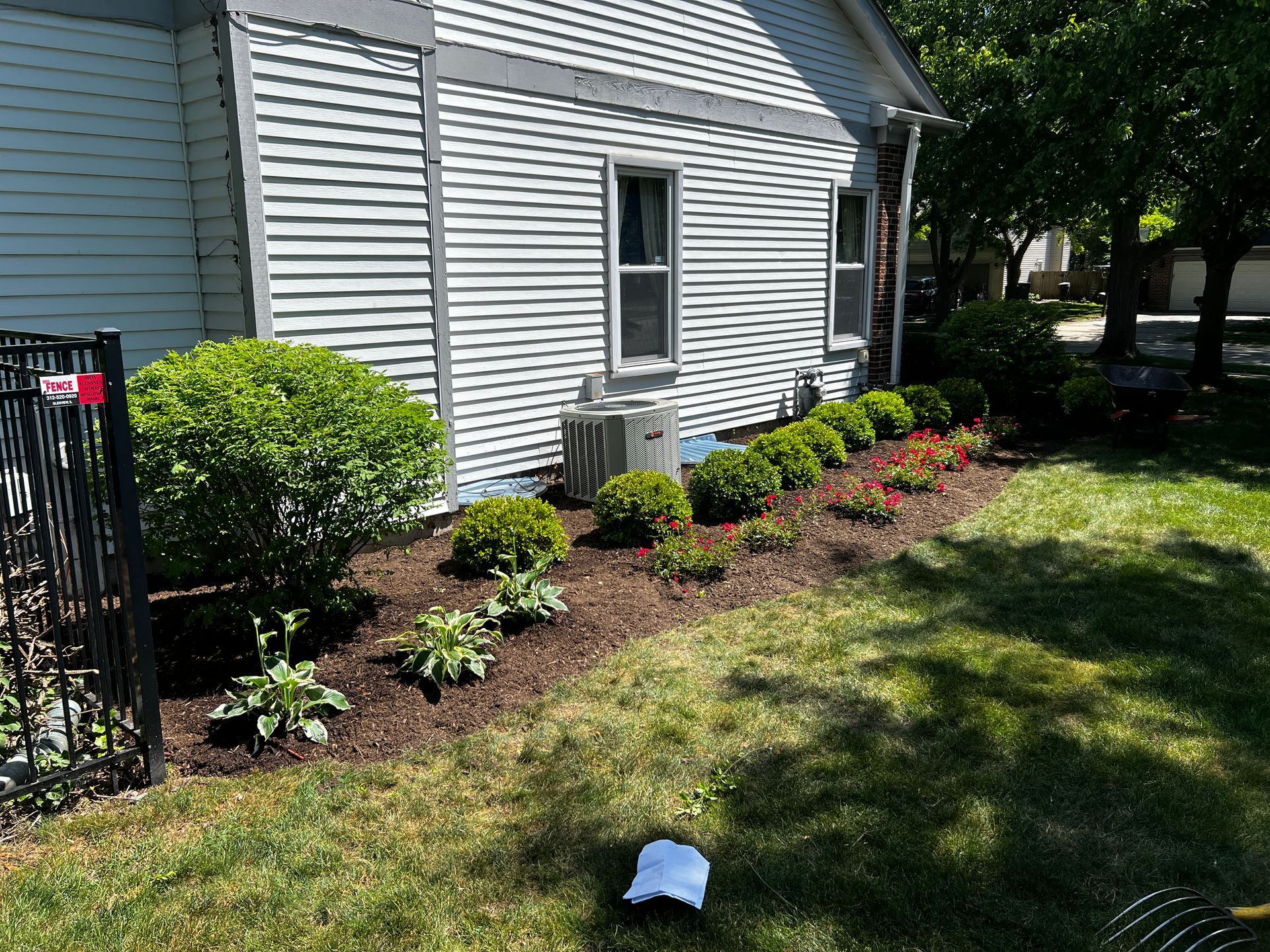 Front yard flower bed along white house siding with shrubs, mulch, and a green lawn in sunlight