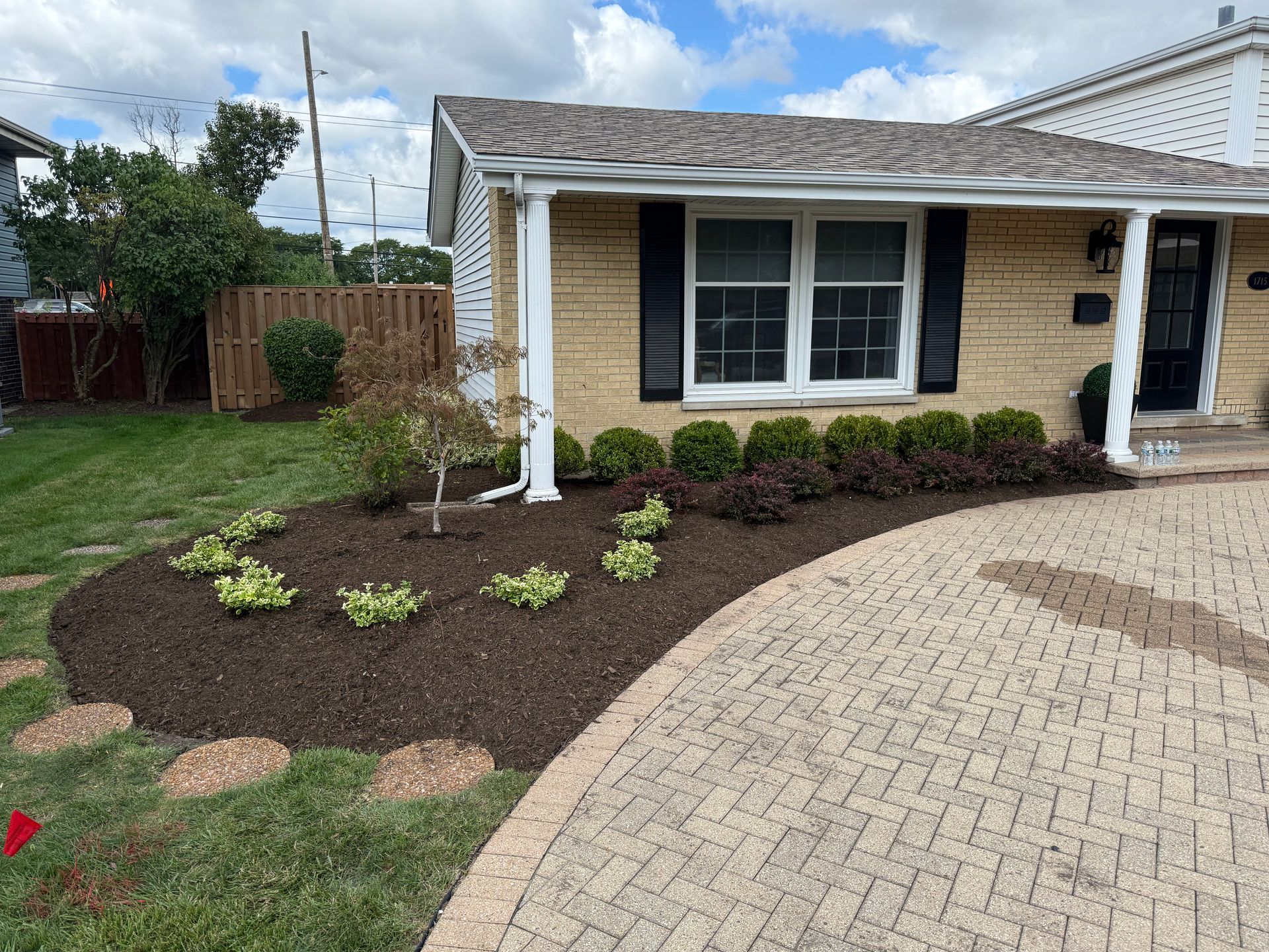 Front yard landscaping beside a beige house with mulch beds, small shrubs, and a curved brick driveway.