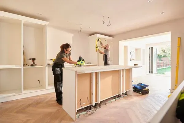 Two people installing kitchen cabinets with tools, working inside.