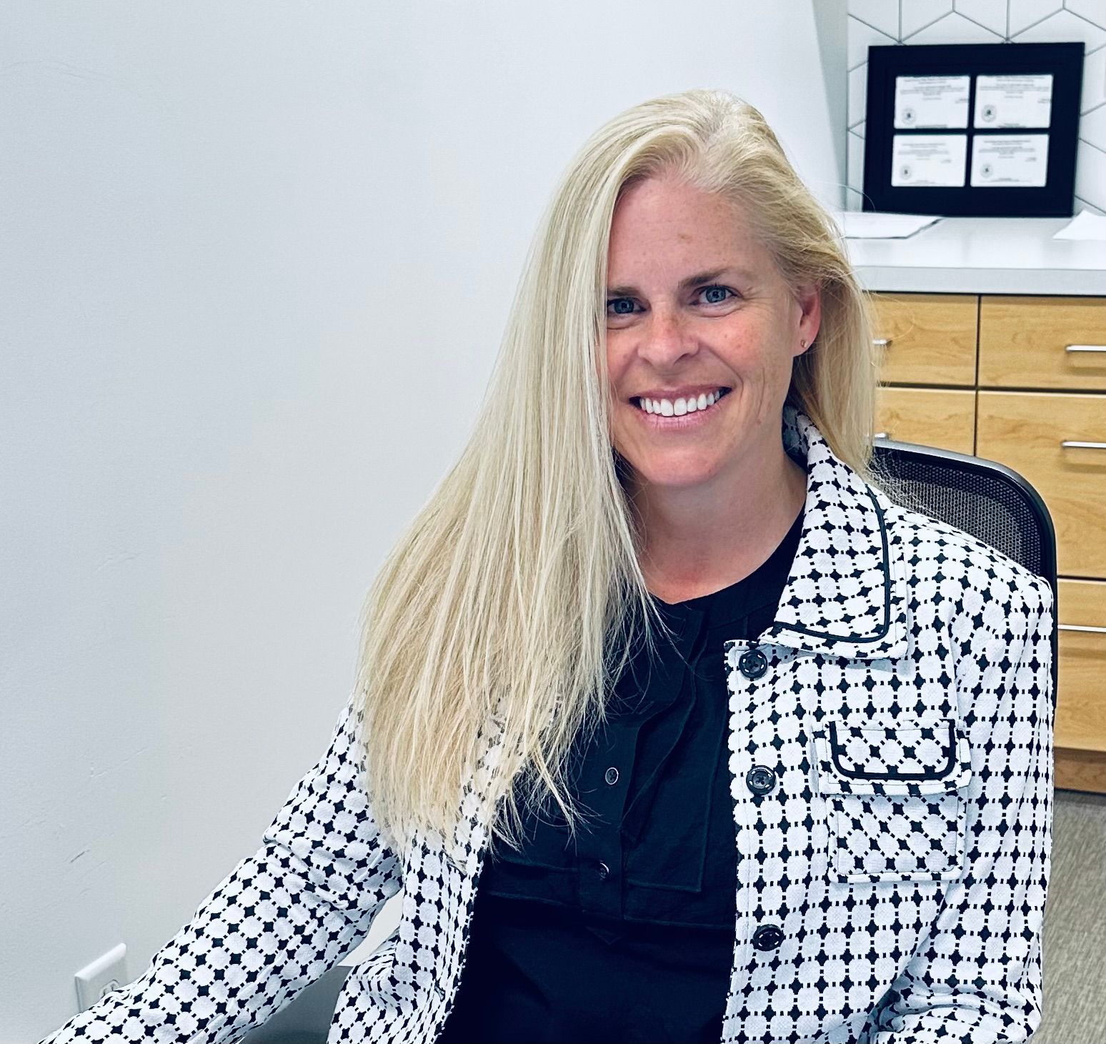 A woman in a black and white jacket is smiling while sitting in a chair.