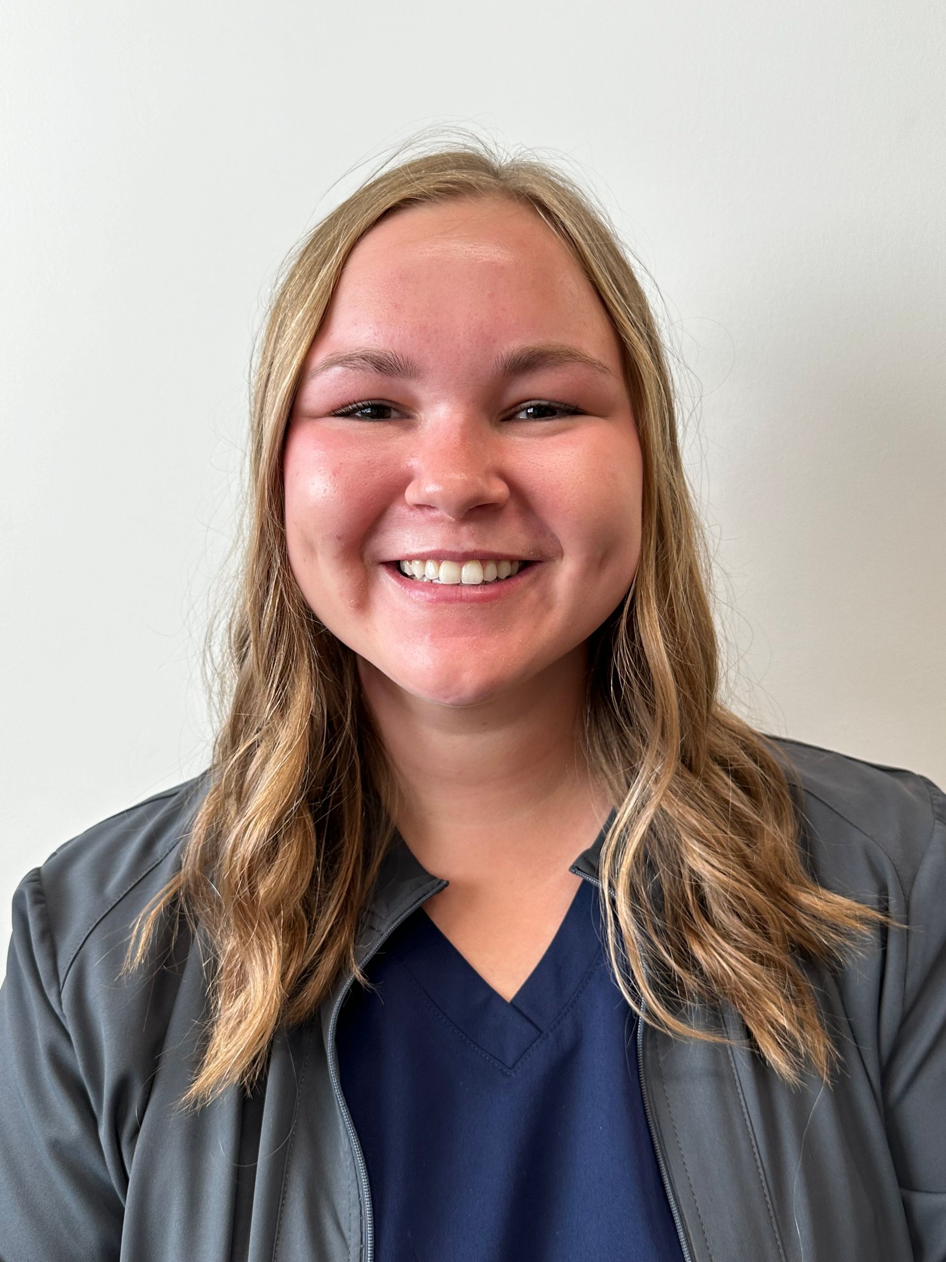 A woman wearing a gray jacket and a blue scrub top is smiling for the camera.