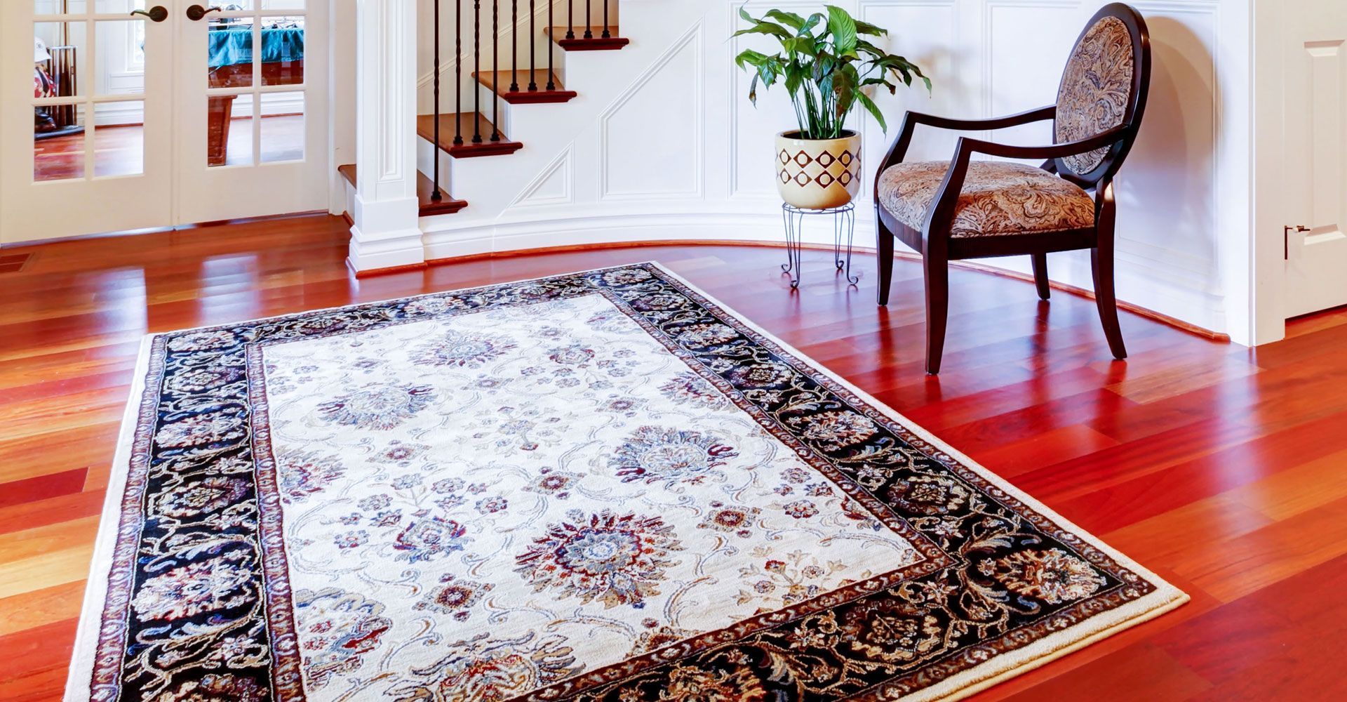 A patterned rug on reddish hardwood floor, a chair, and a plant next to a staircase.