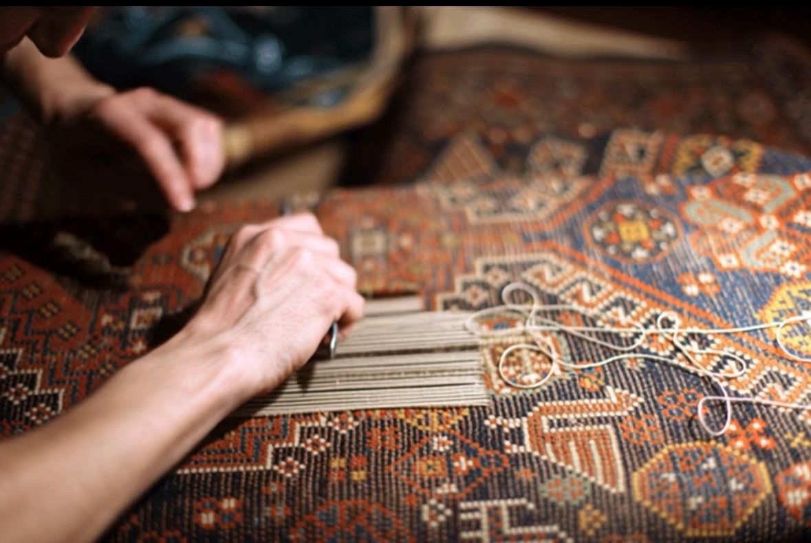 Hands weaving threads into an ornate rug; close-up shot, brown and blue colors.