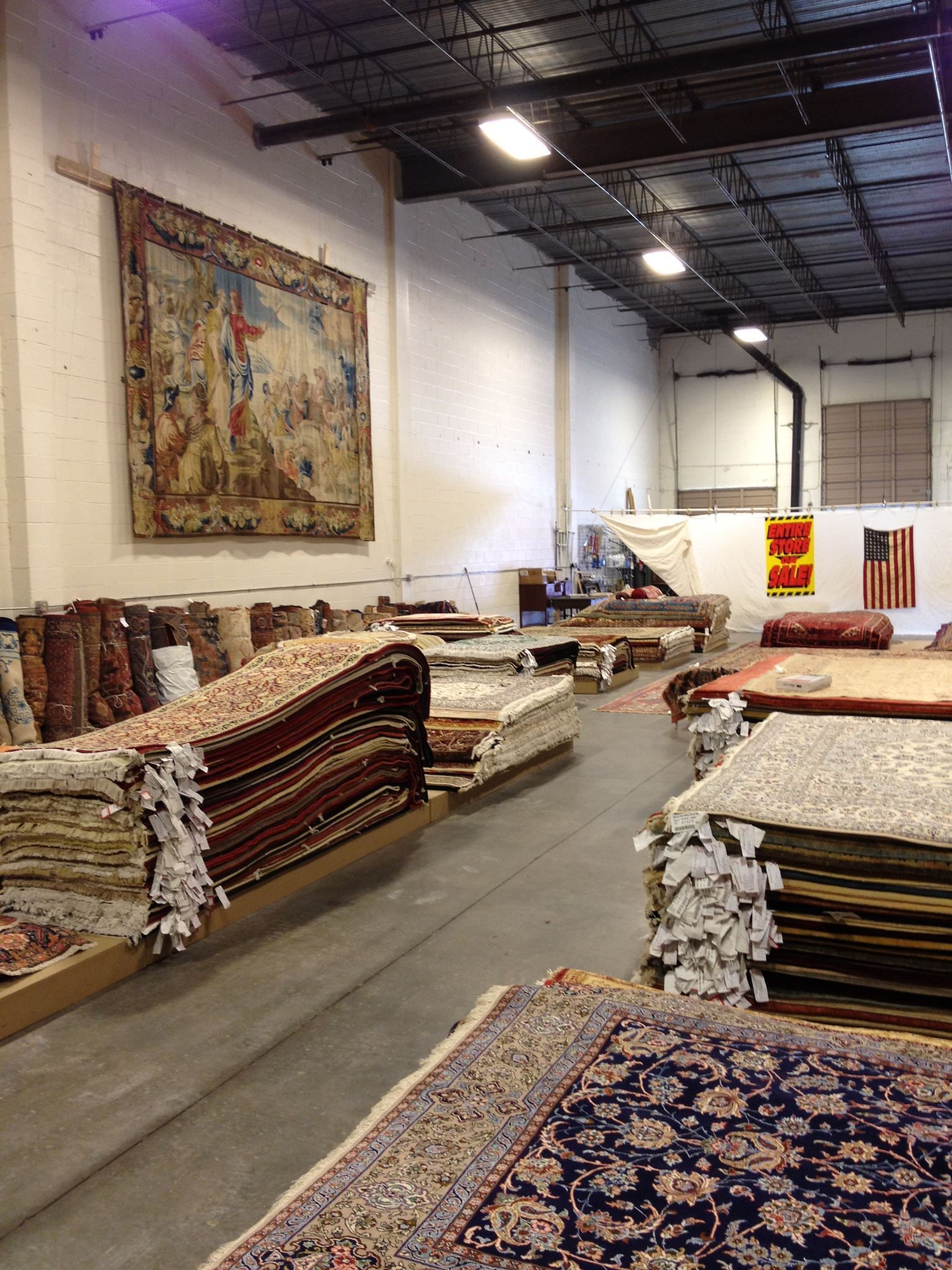 Warehouse with stacks of rugs; tapestry on wall, overhead lights.