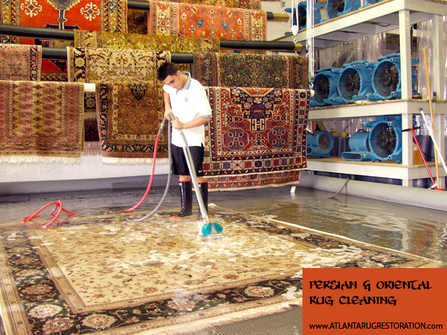Man cleaning a rug with a hose in a rug cleaning facility. Other rugs are hanging.