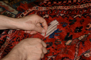 Hands repairing a red and blue patterned rug, weaving threads into a damaged area.