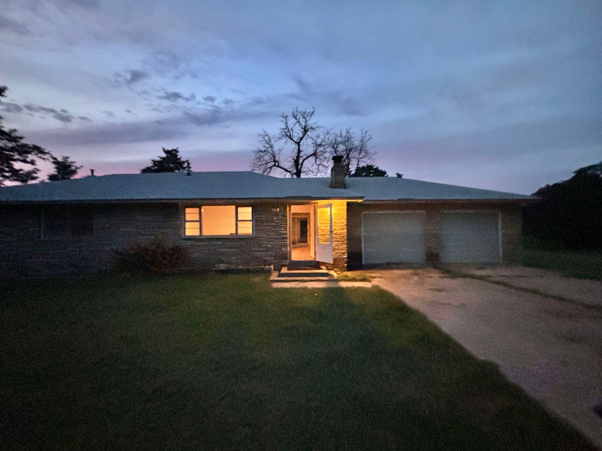 House exterior at dusk, with lit windows and garage. Overcast sky, long driveway, and lawn.