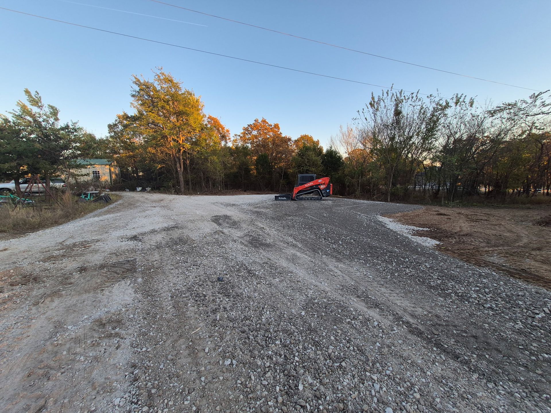 Gravel driveway under construction; orange skid steer on the right. Trees and blue sky in the background.