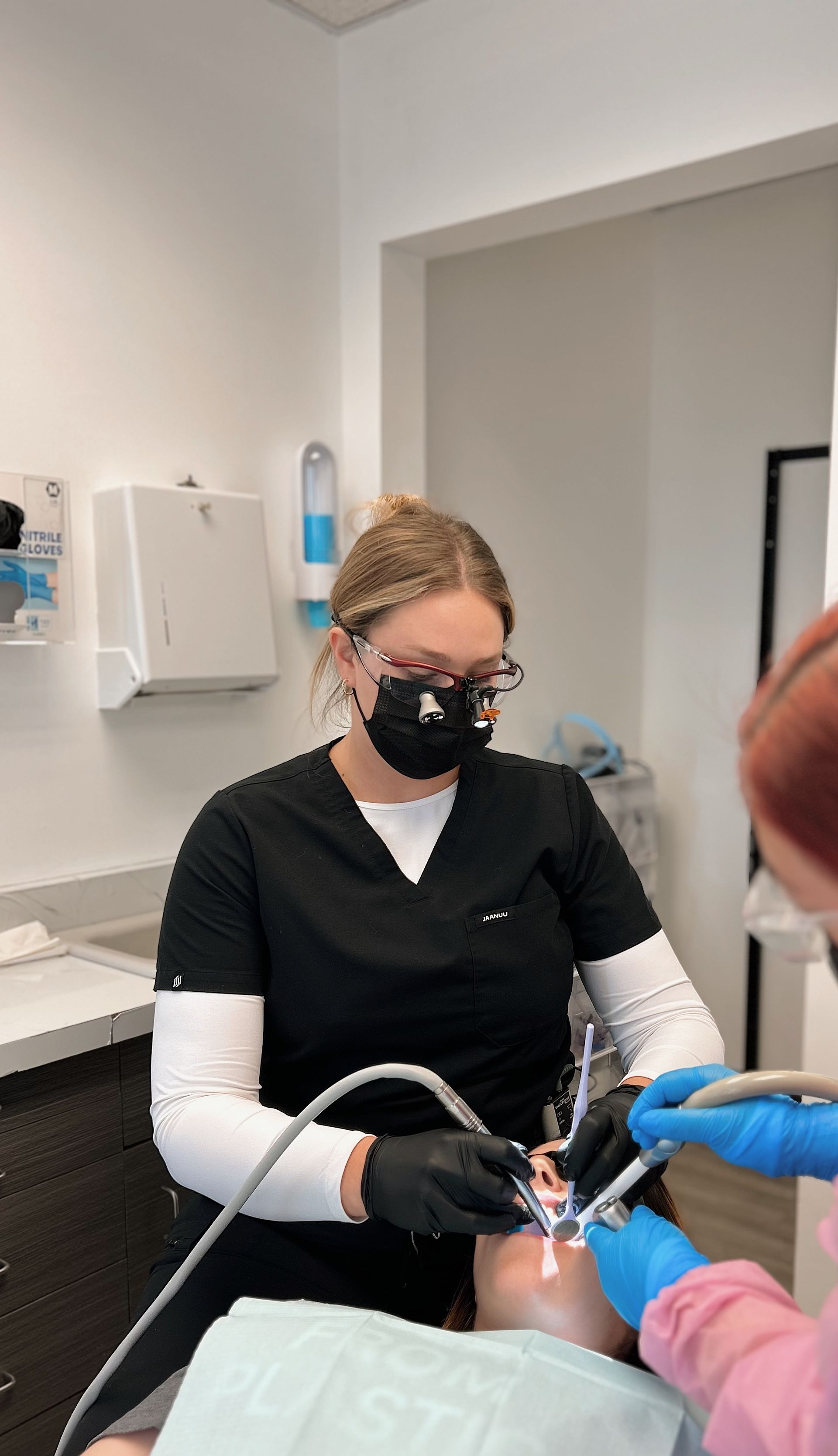 Dentist wearing black scrubs and mask working on a patient's teeth in a dental office.