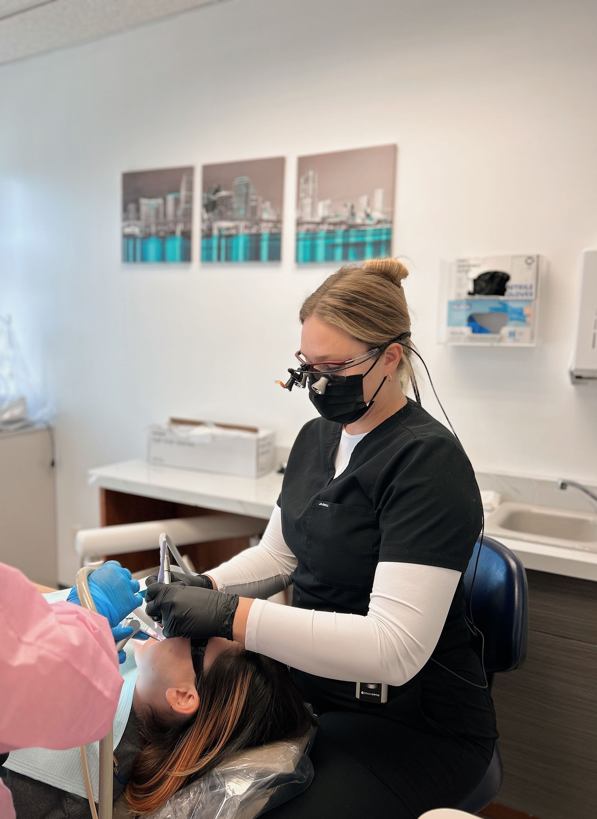 Dentist in black scrubs examining a patient. Surgical mask, loupes, and black gloves. Dental office setting.