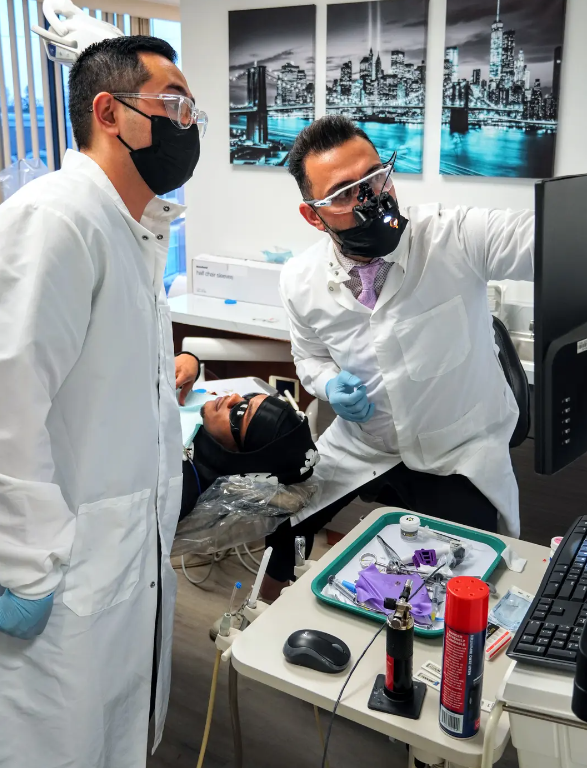 Two dentists wearing masks and goggles are talking to a patient.