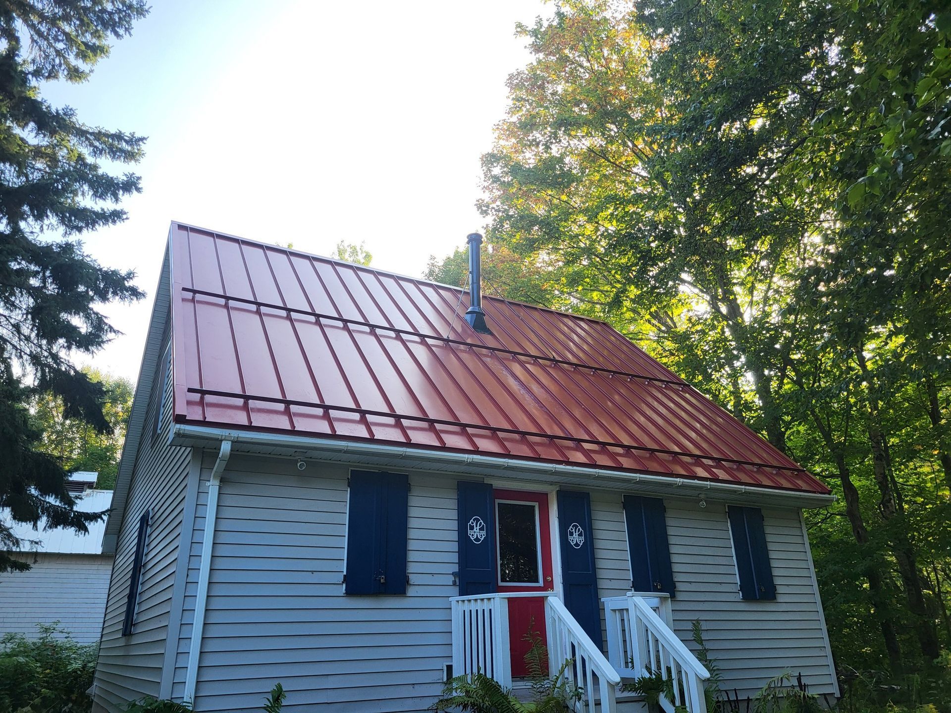 Small gray house with red metal roof, blue shutters, and red door, surrounded by trees.