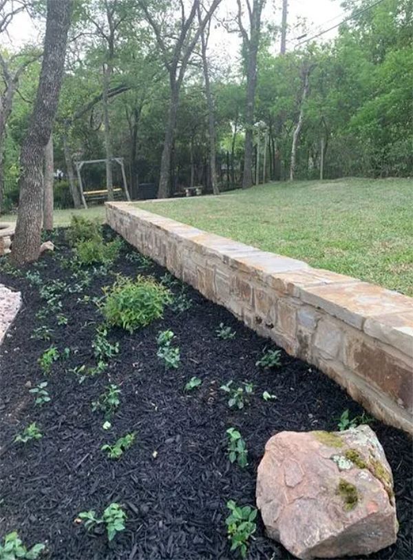 Stone retaining wall beside a landscaped garden bed with mulch, small plants, and trees in a wooded yard
