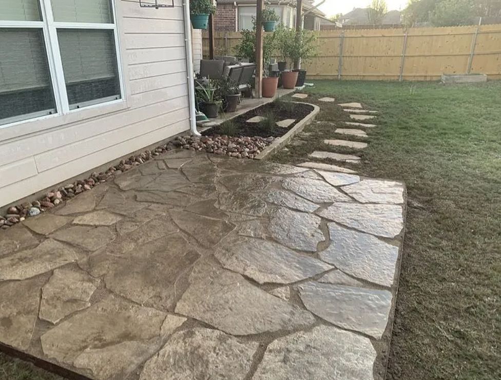 Stone patio beside a house, leading to a grassy backyard and fenced yard.