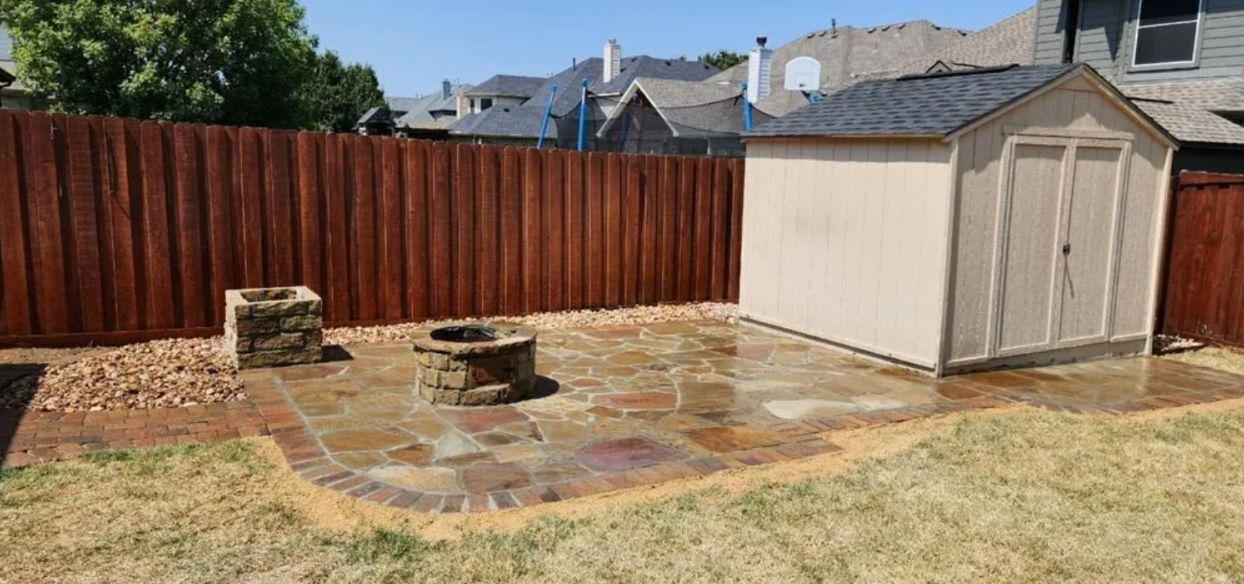 Backyard with wooden fence, stone patio, fire pit, and a beige storage shed under a bright sky