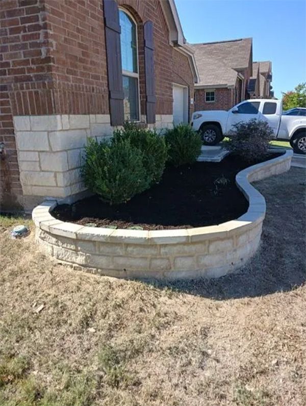 Curved stone garden bed with shrubs beside a brick house and driveway, with a white truck in the background