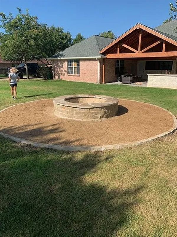 Backyard patio with a round stone fire pit and a covered brick house; person walking on the lawn.