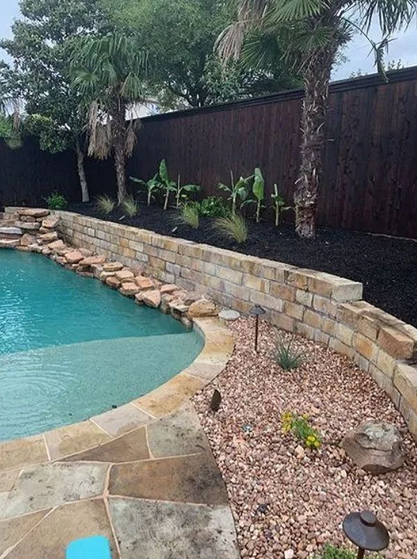 Backyard pool with stone coping, rock border, tropical plants, and a dark wood fence