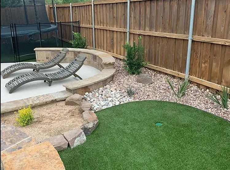 Backyard patio with lounge chairs, artificial grass, rock border, and wooden fence
