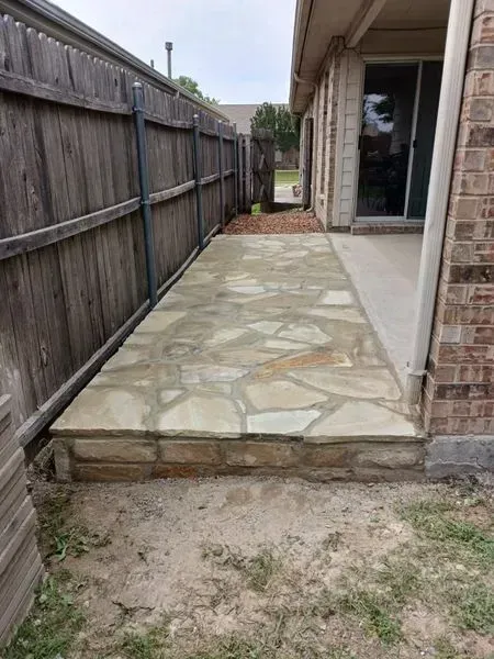 Narrow stone-paved side yard between a wooden fence and a brick house, leading to a gate.