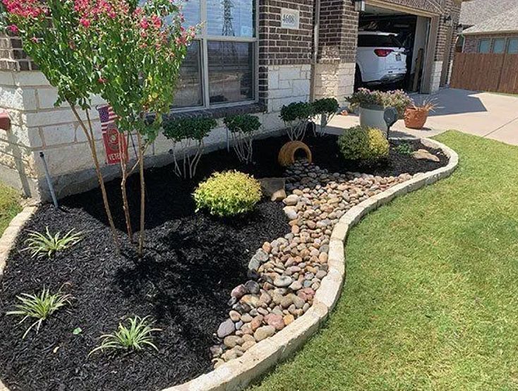 Curved flower bed with black mulch, rocks, small plants, and a lawn beside a brick house