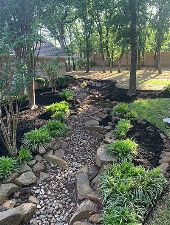 Stone-lined dry creek bed winding through a landscaped backyard with trees and shrubs.