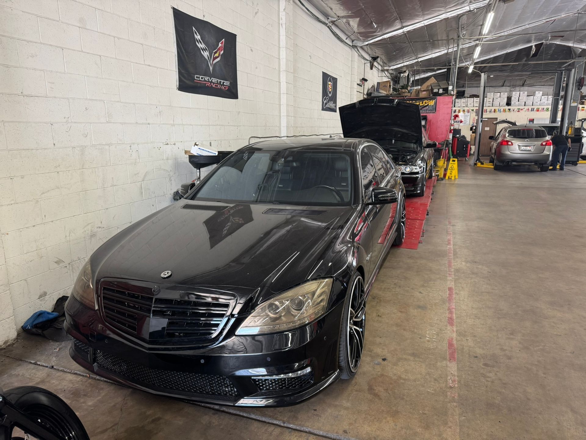 Black sedan in an auto shop, parked beside tires and tools under bright overhead lights.