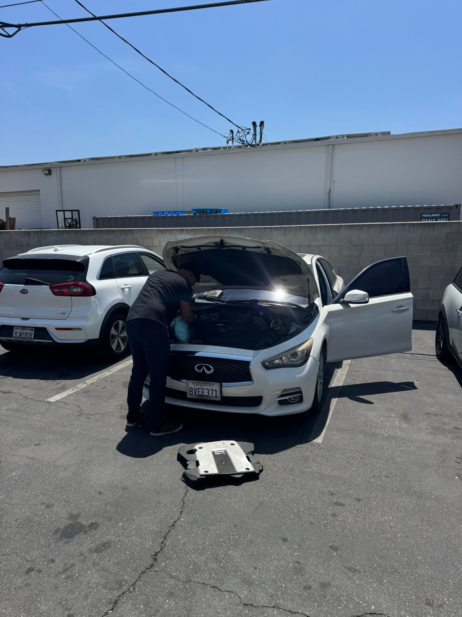 Person works under the hood of a white car in a parking lot; another white SUV is parked beside it.