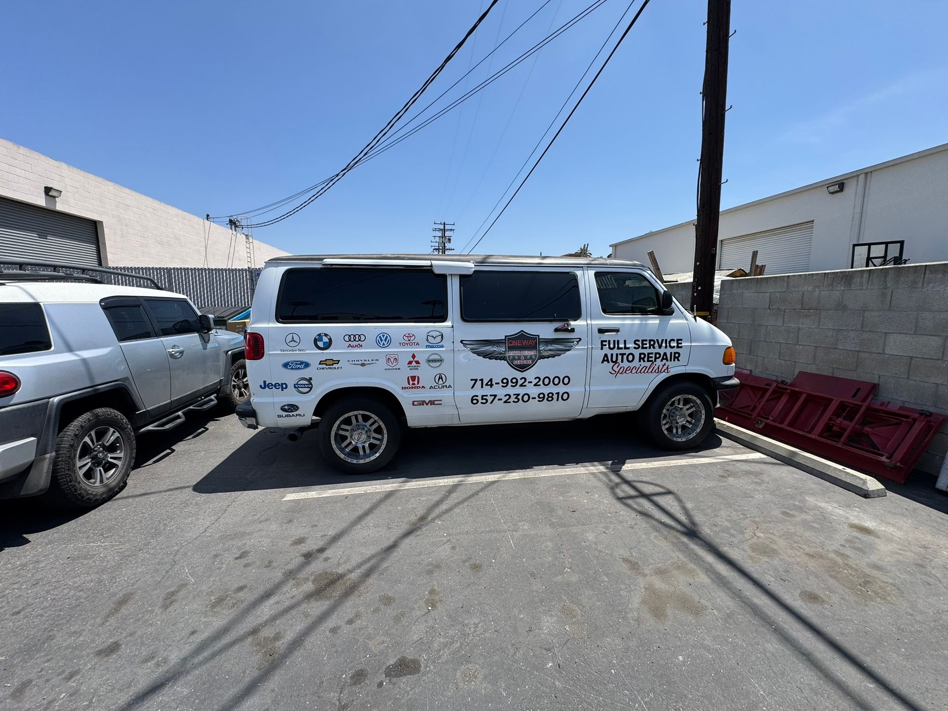 White cargo van with company logos parked in an industrial lot beside other vehicles and red pallets.