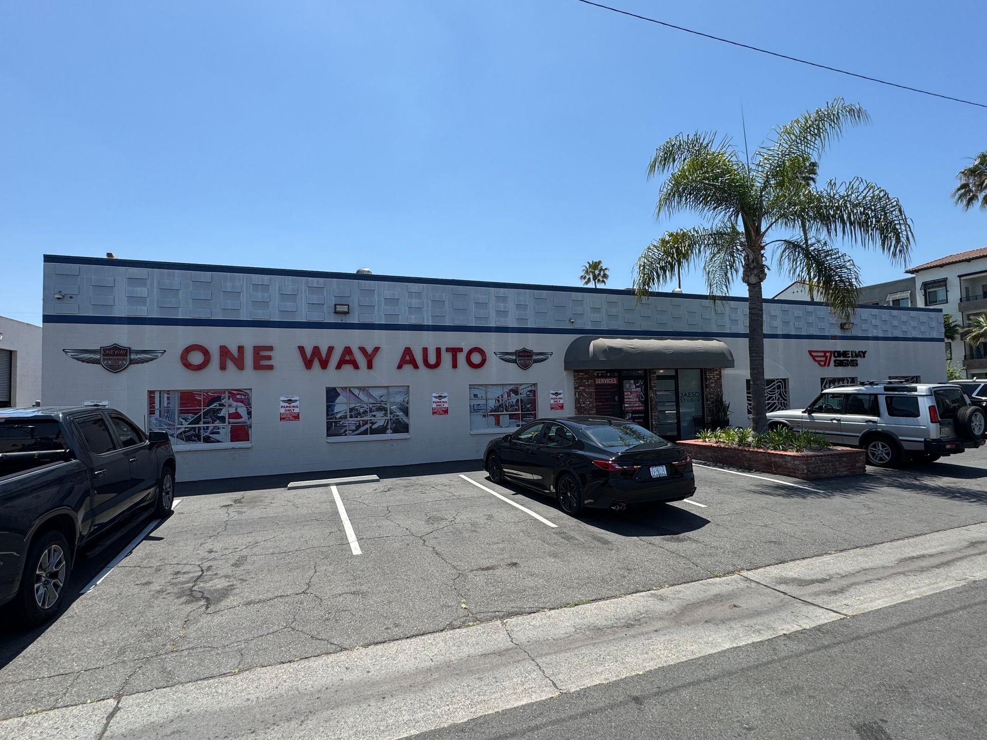 One Way Auto storefront with parked cars in a sunny lot, palm trees, and a blue sky