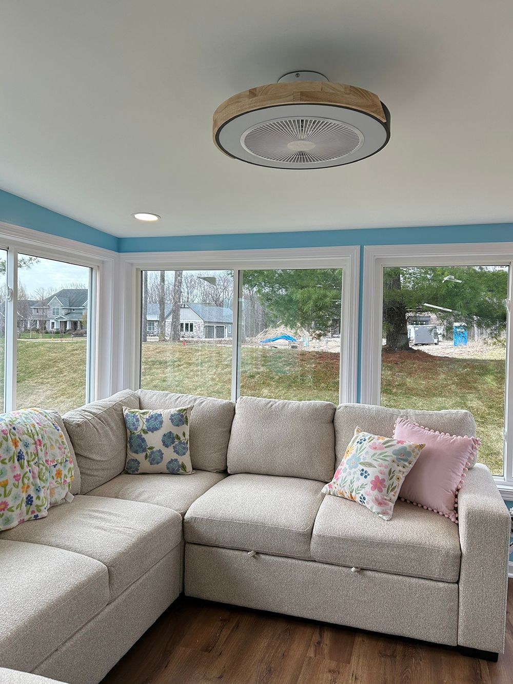 A light beige sectional sofa with patterned throw pillows sits in a sunroom with windows overlooking a grassy yard.