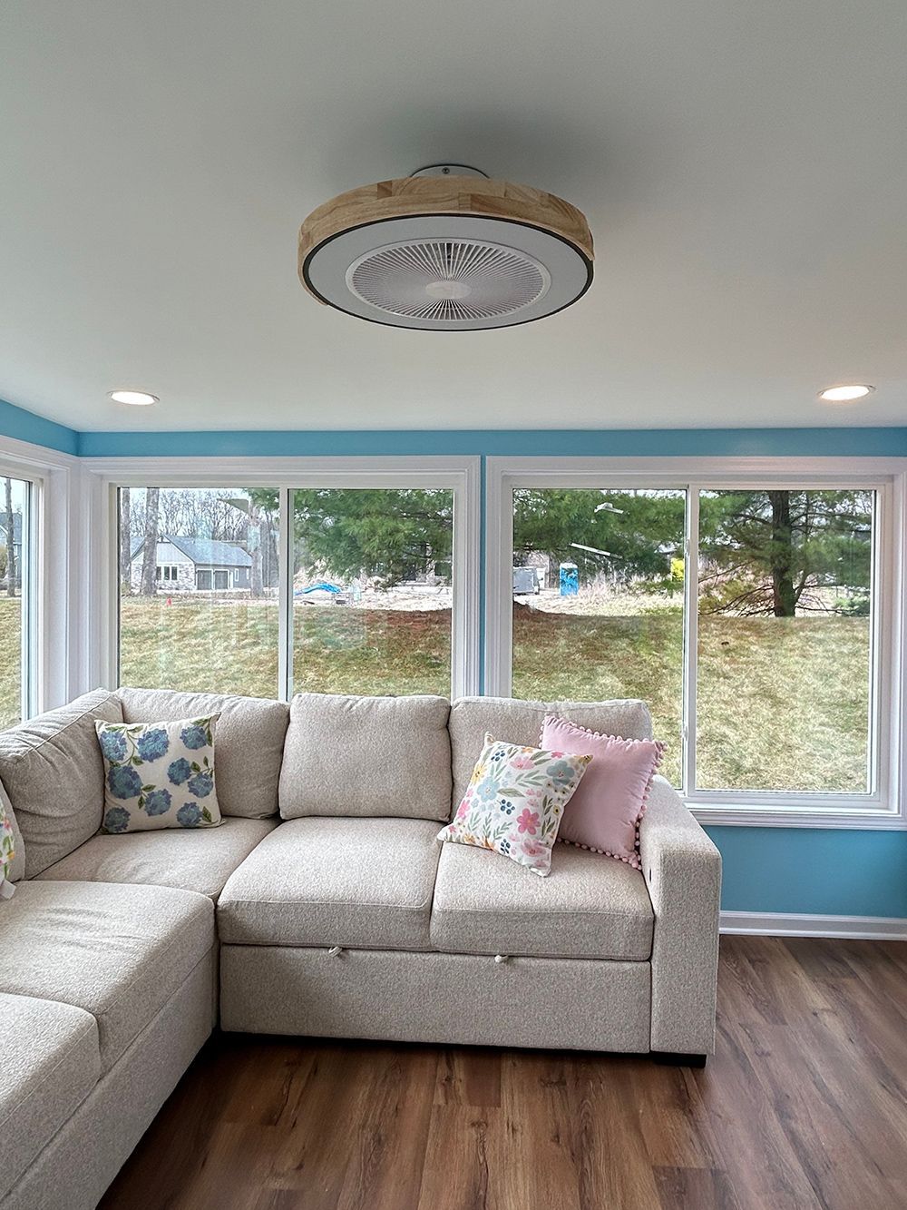 A light-colored sectional sofa sits in a sunroom with blue walls, large windows, and a circular ceiling fan light fixture.