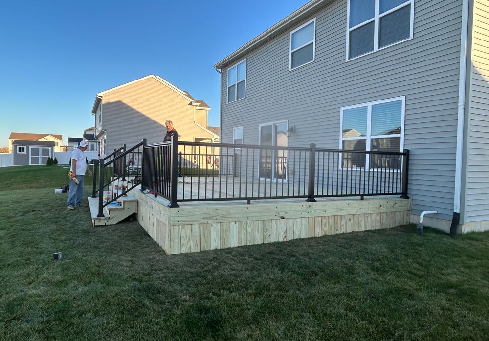 Two people stand on a newly constructed wooden deck with black metal railings attached to the back of a house.