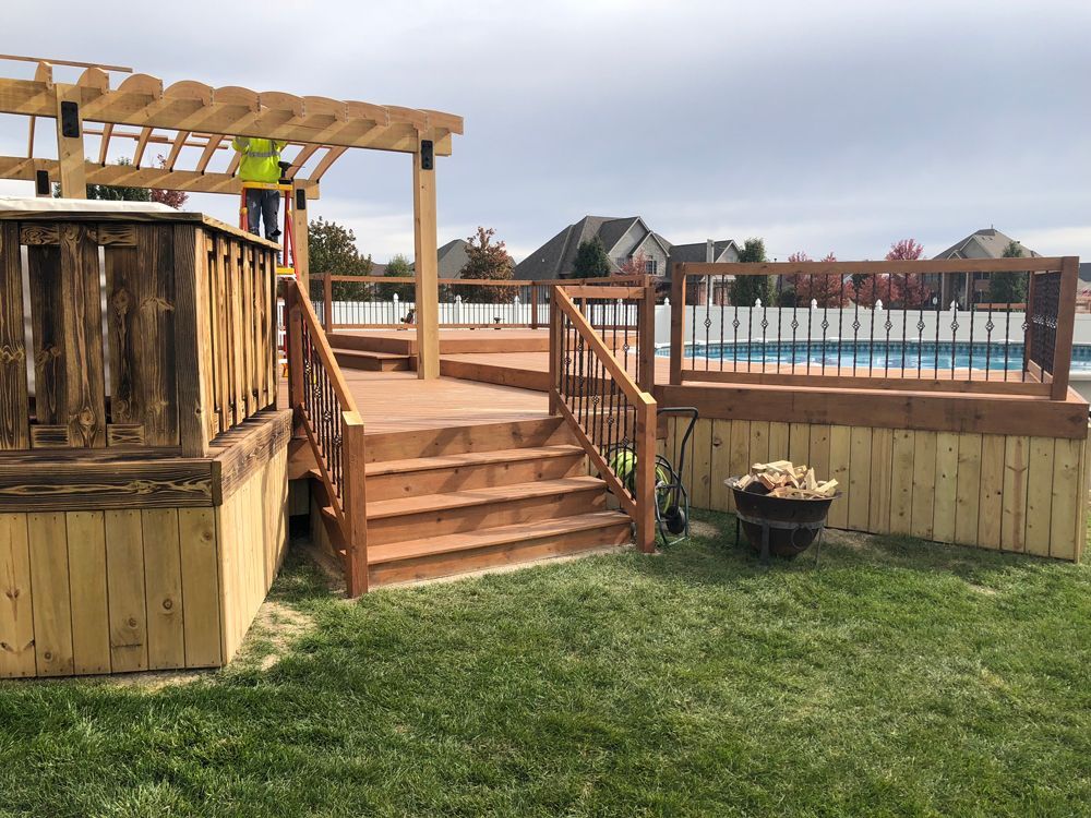A person on a ladder builds a pergola over a wooden deck with stairs and a pool in the background, near a fire pit.