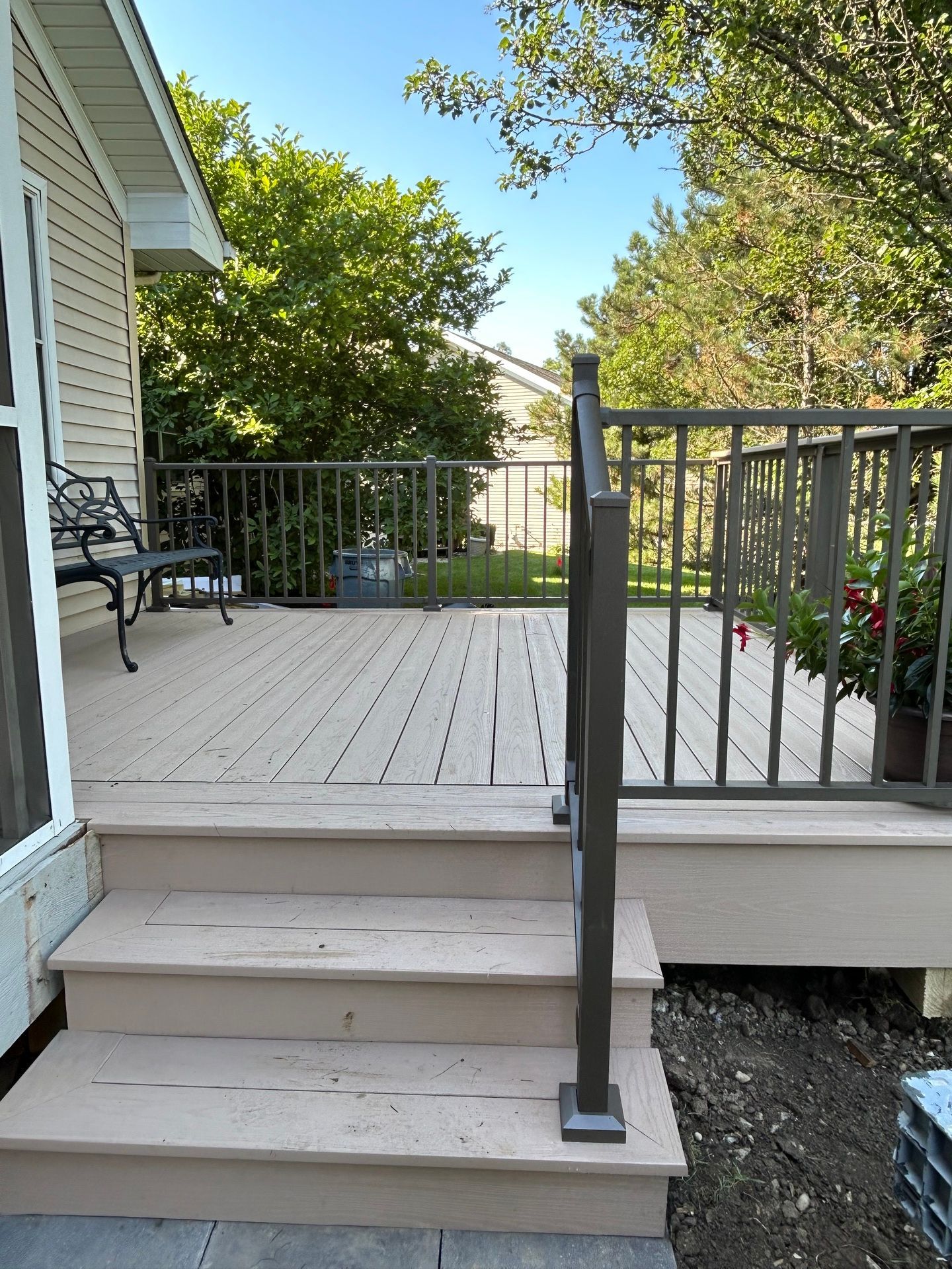 Porch steps with a black railing leading to a light gray deck beside a house and garden.