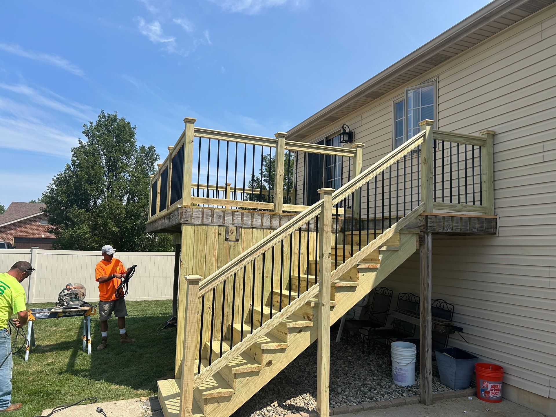 A worker in an orange shirt uses a power tool on a wooden deck staircase under construction next to a house.