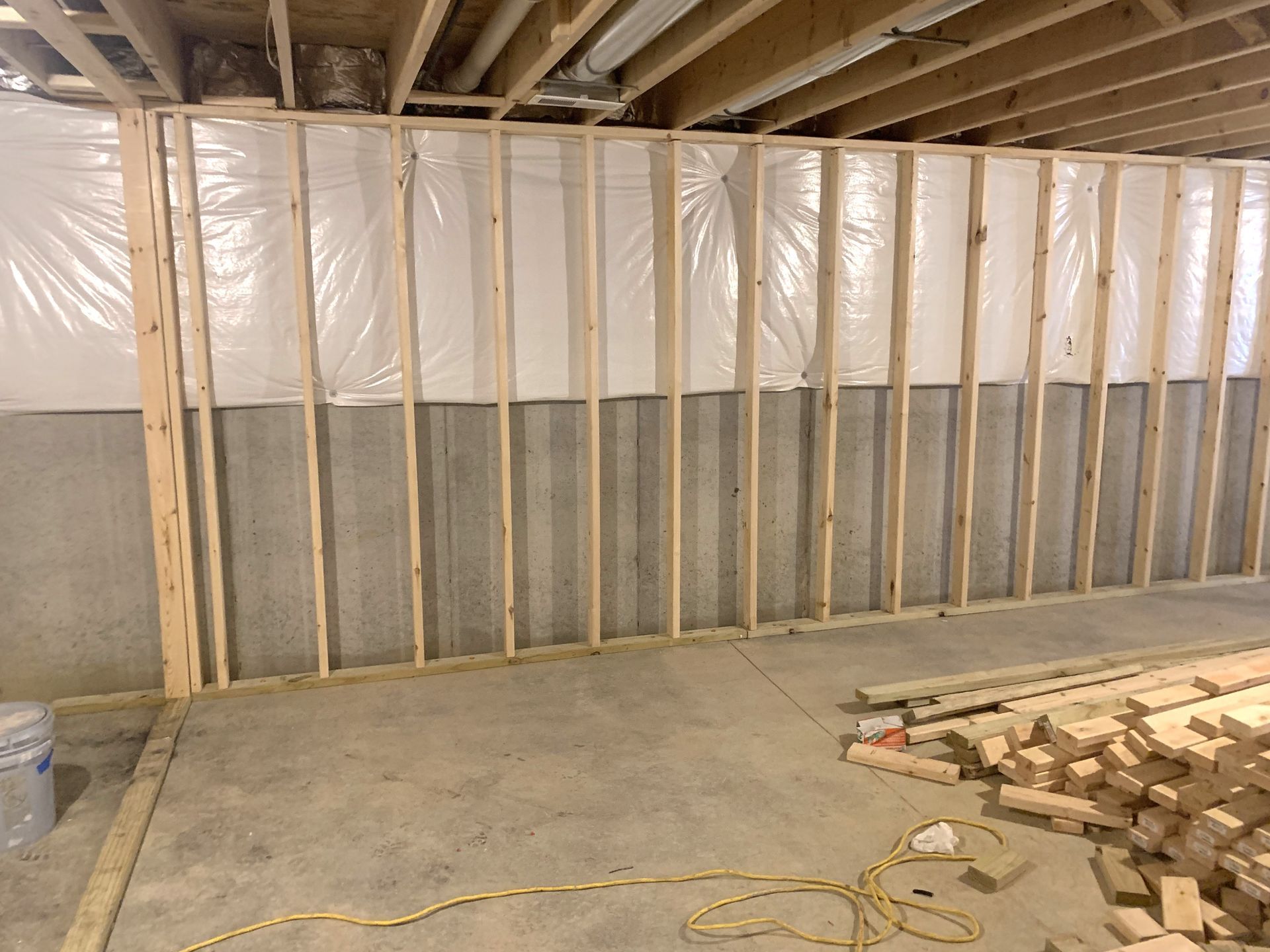 Basement wall under construction: wooden frame, insulation, concrete, wood piles, and electrical cord on the floor.