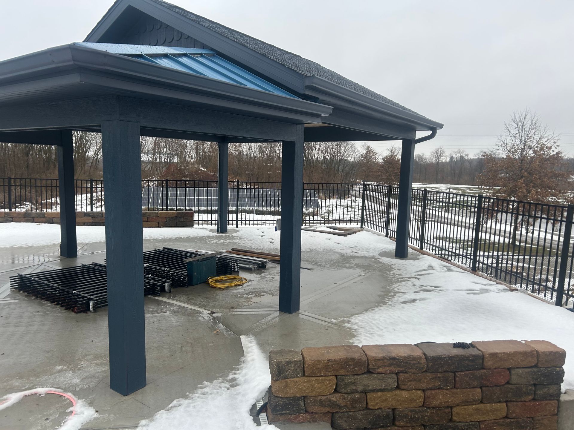 Gazebo with dark blue columns and roof on a concrete patio with snow and a brick wall. Black fence in background.
