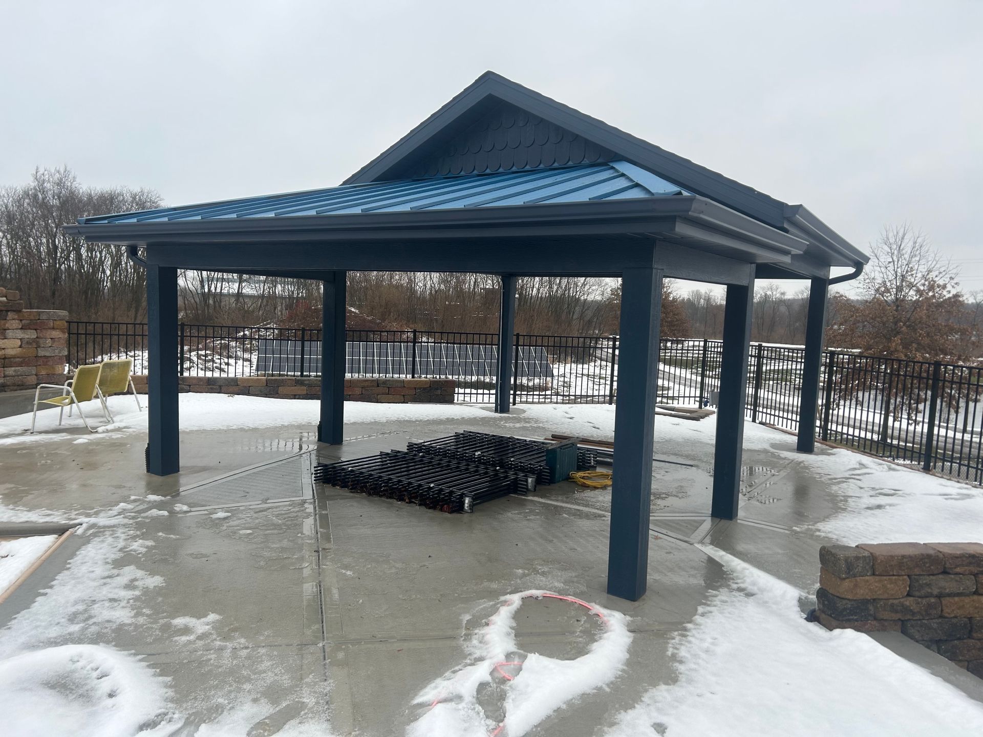Gray gazebo with blue roof on a snowy concrete patio, overlooking a black metal fence and trees.
