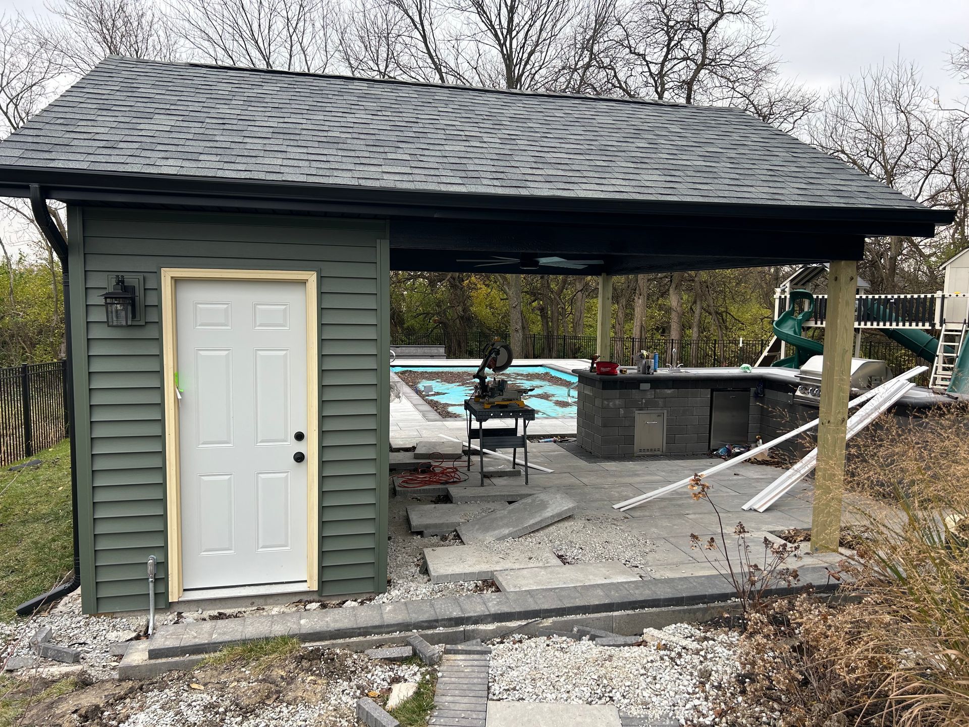 Green shed with white door and attached covered patio next to a pool.