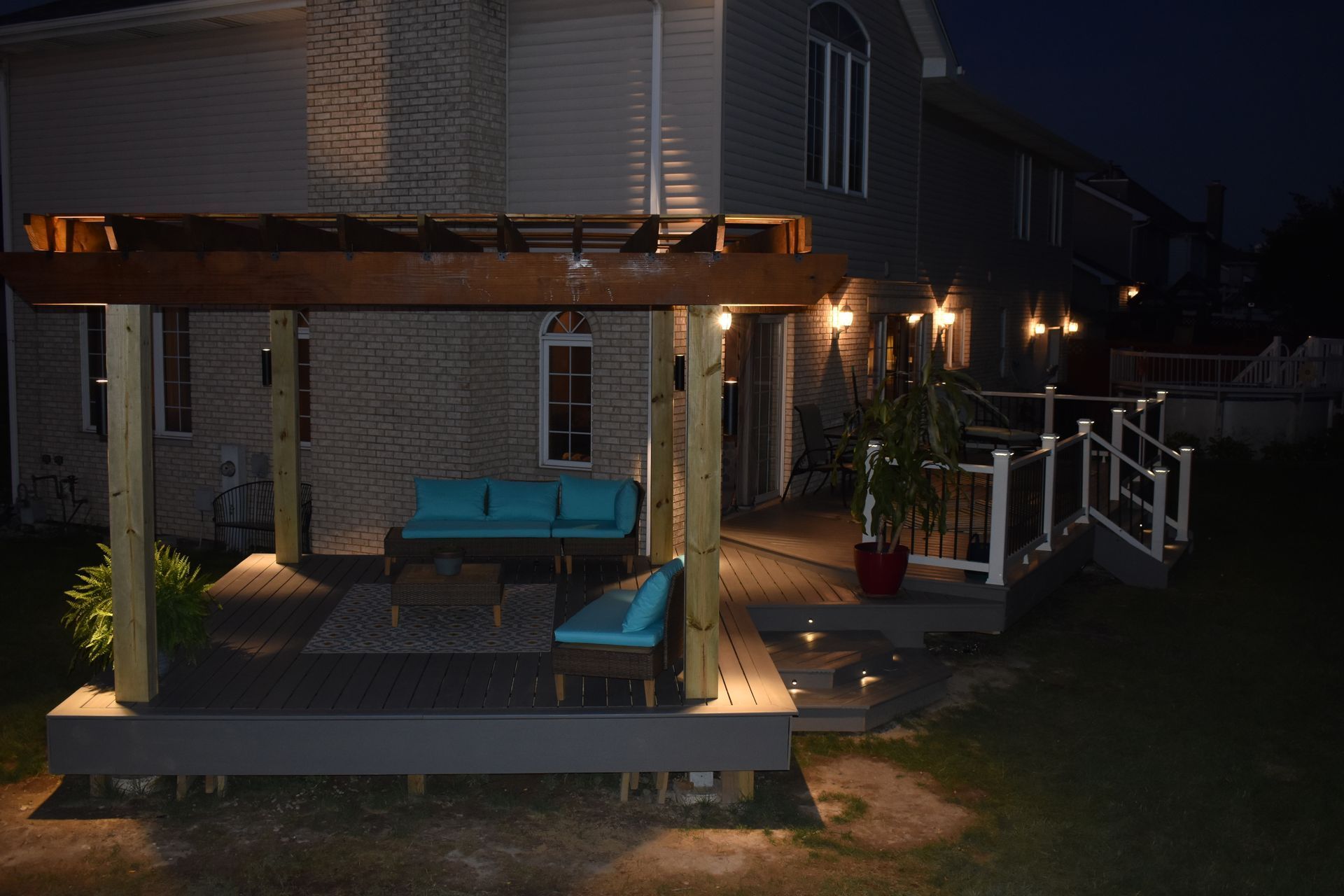 Outdoor deck with pergola, illuminated at night. Furnished with teal seating, surrounded by ambient lighting.
