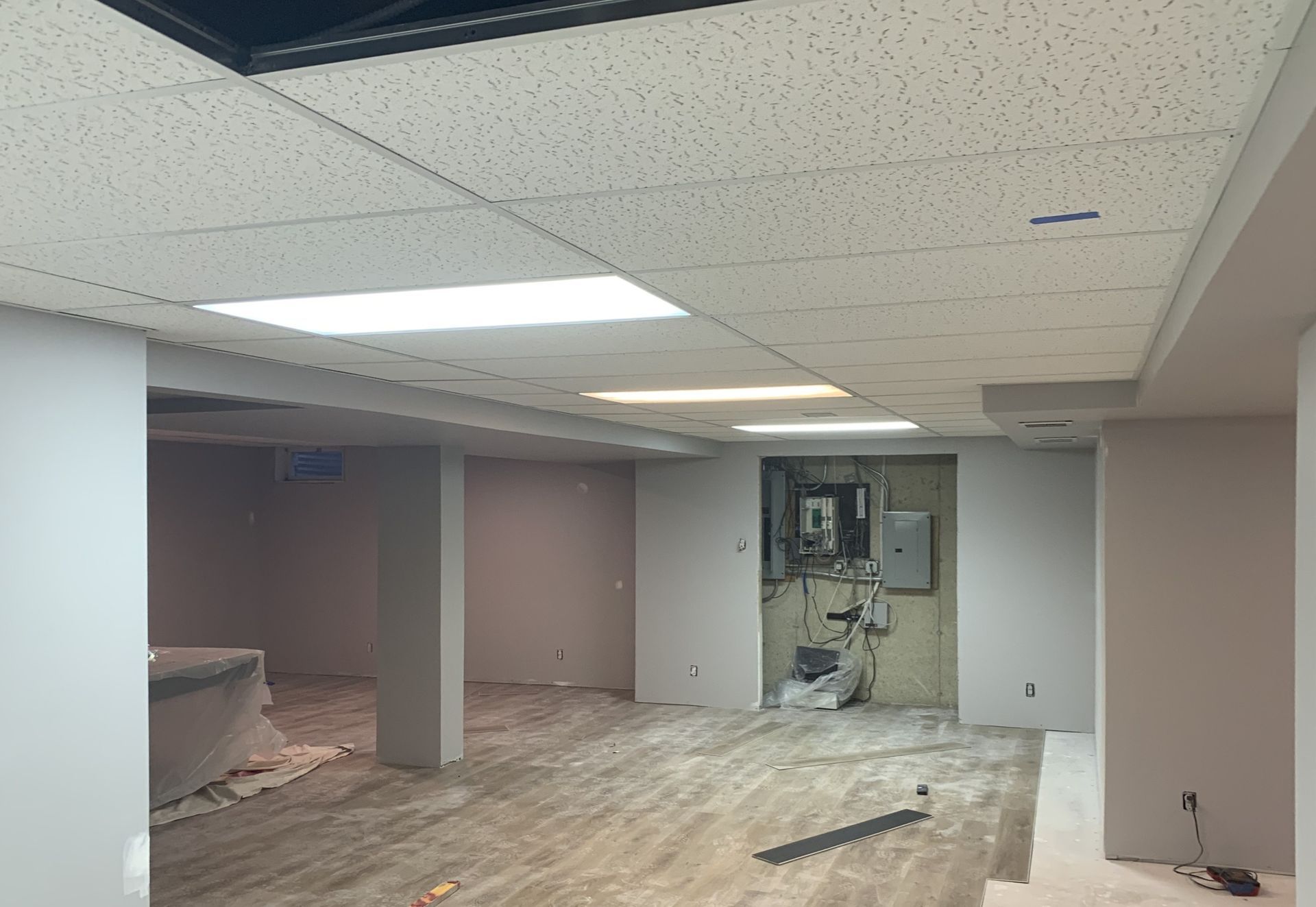 Room with unfinished construction: exposed ceiling tiles, bare floors, and electrical panel.