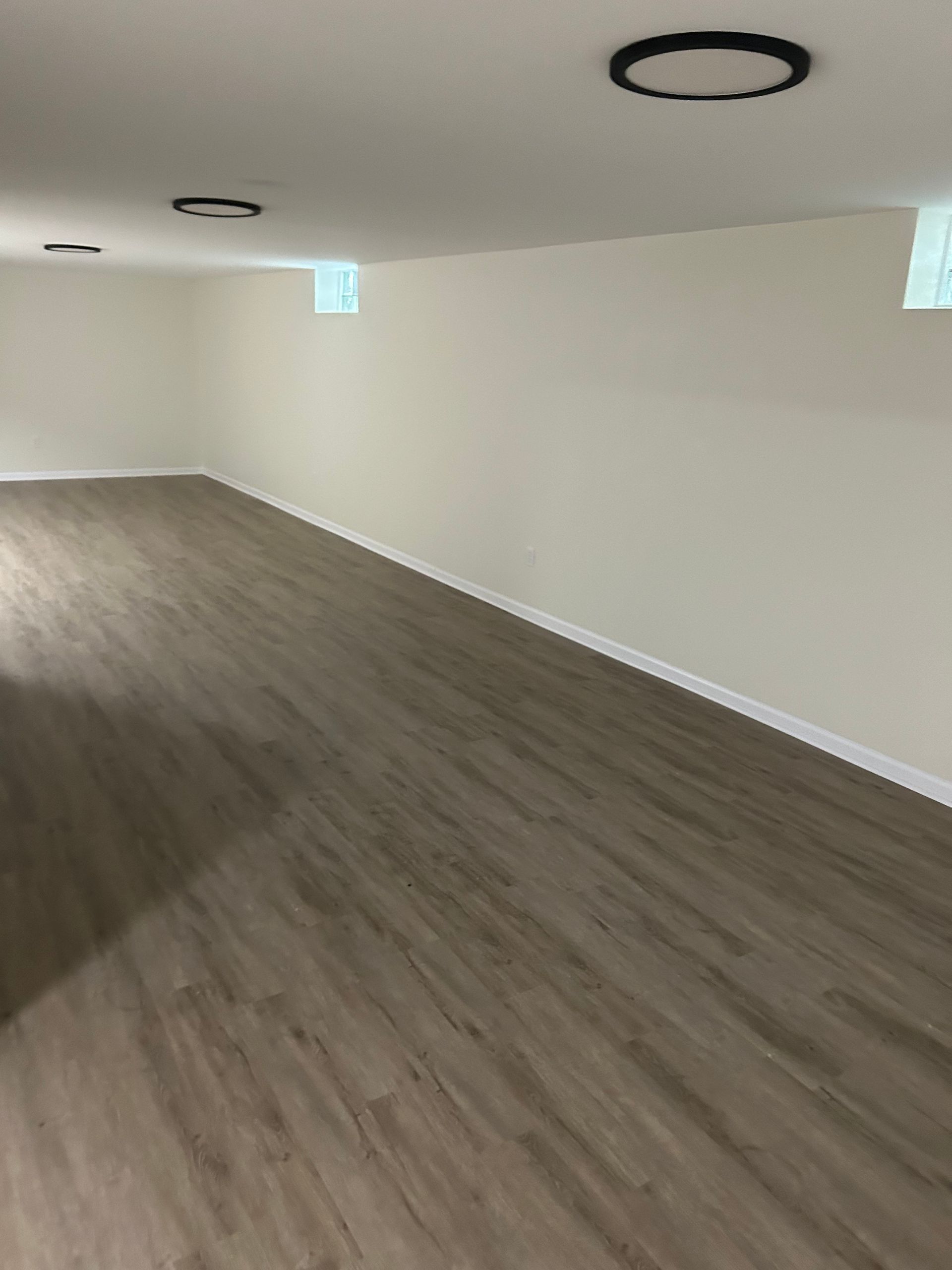 Finished basement with light wood-look flooring, beige walls, recessed lighting, and small square windows.