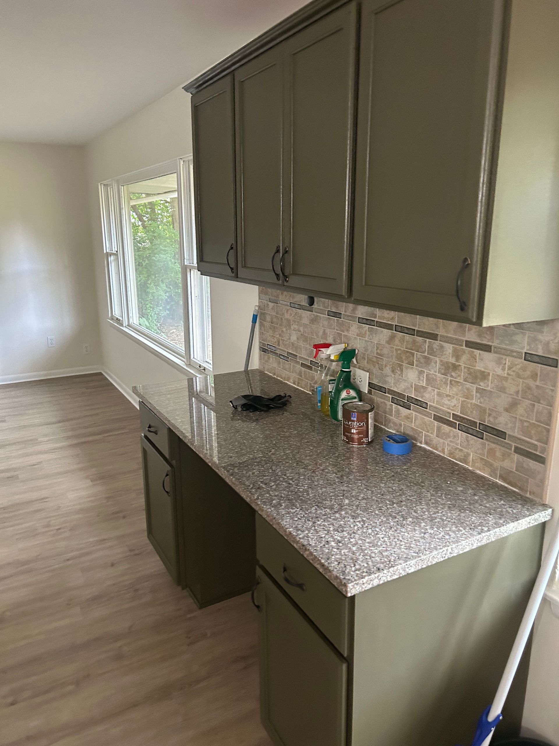 Olive green kitchen cabinets and countertop, neutral backsplash, window in the background.
