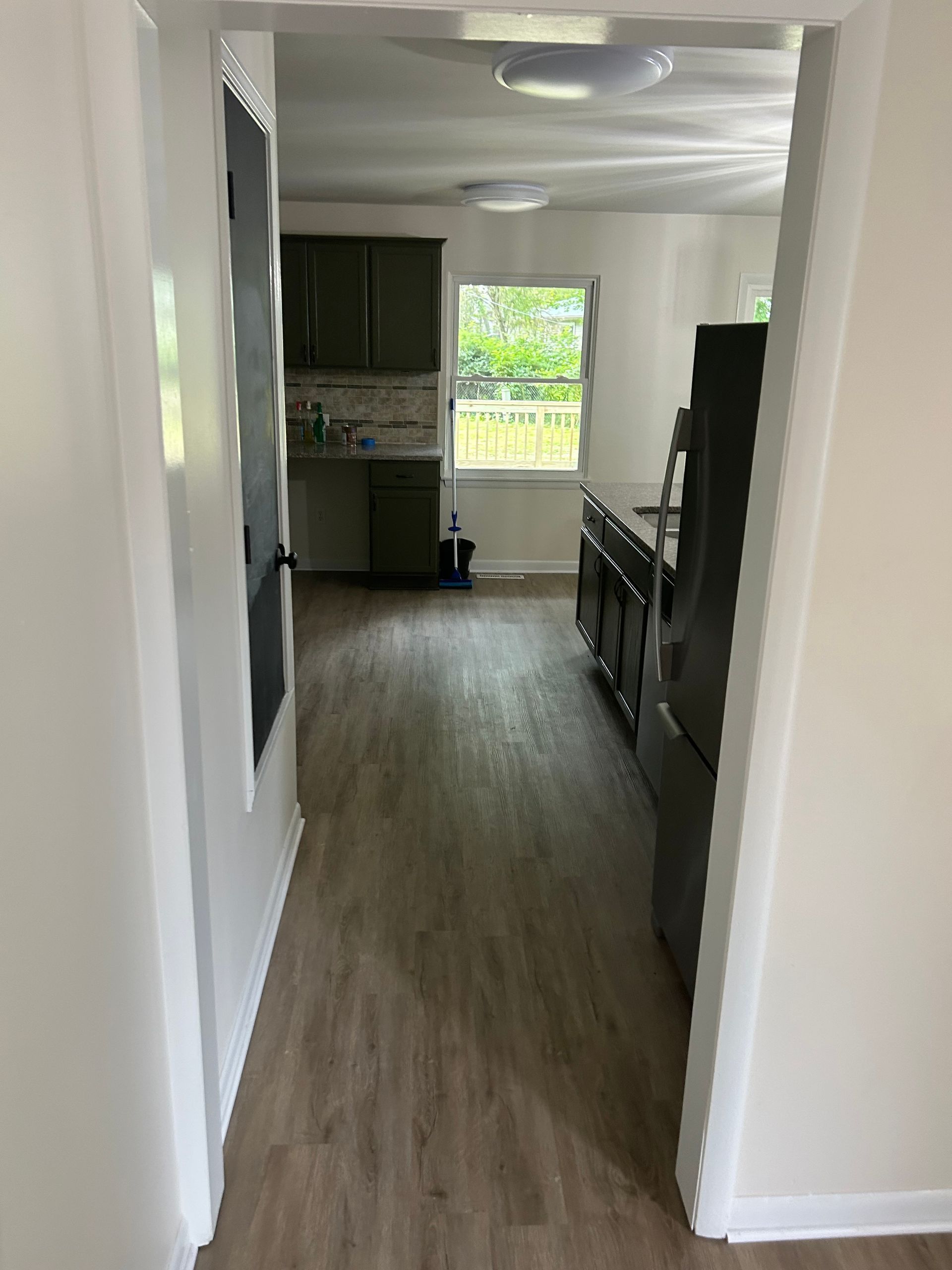 View into a kitchen: olive cabinets, stainless steel fridge, light wood-look floor, window with a green outdoor view.