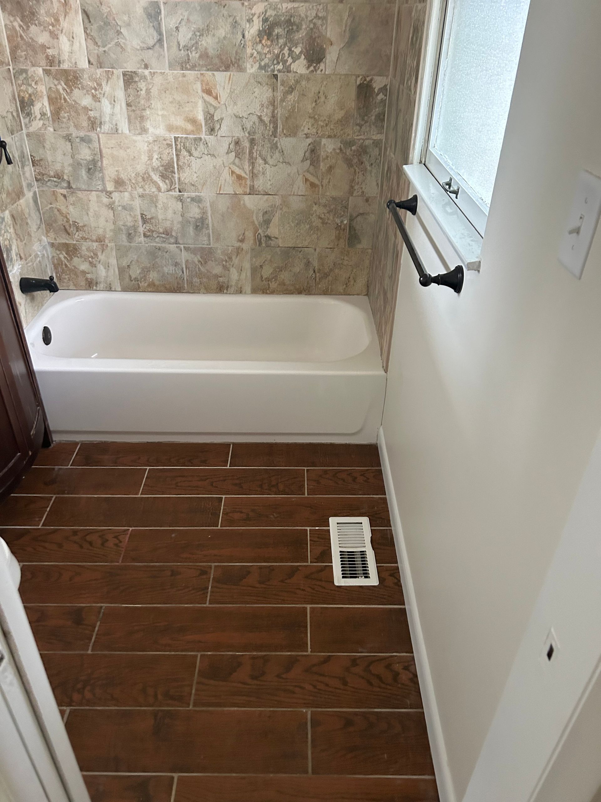 Bathroom with white tub, stone tile wall, wood-look tile floor, window, and towel rack.